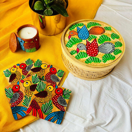 One handwoven bamboo fruit box with a motif of two birds flying over a field, a set of four square wood coasters with a motif of one yellow and one red bird and a double wick scented candle with a bird motif displaced against a yellow background with a plant pot beside them