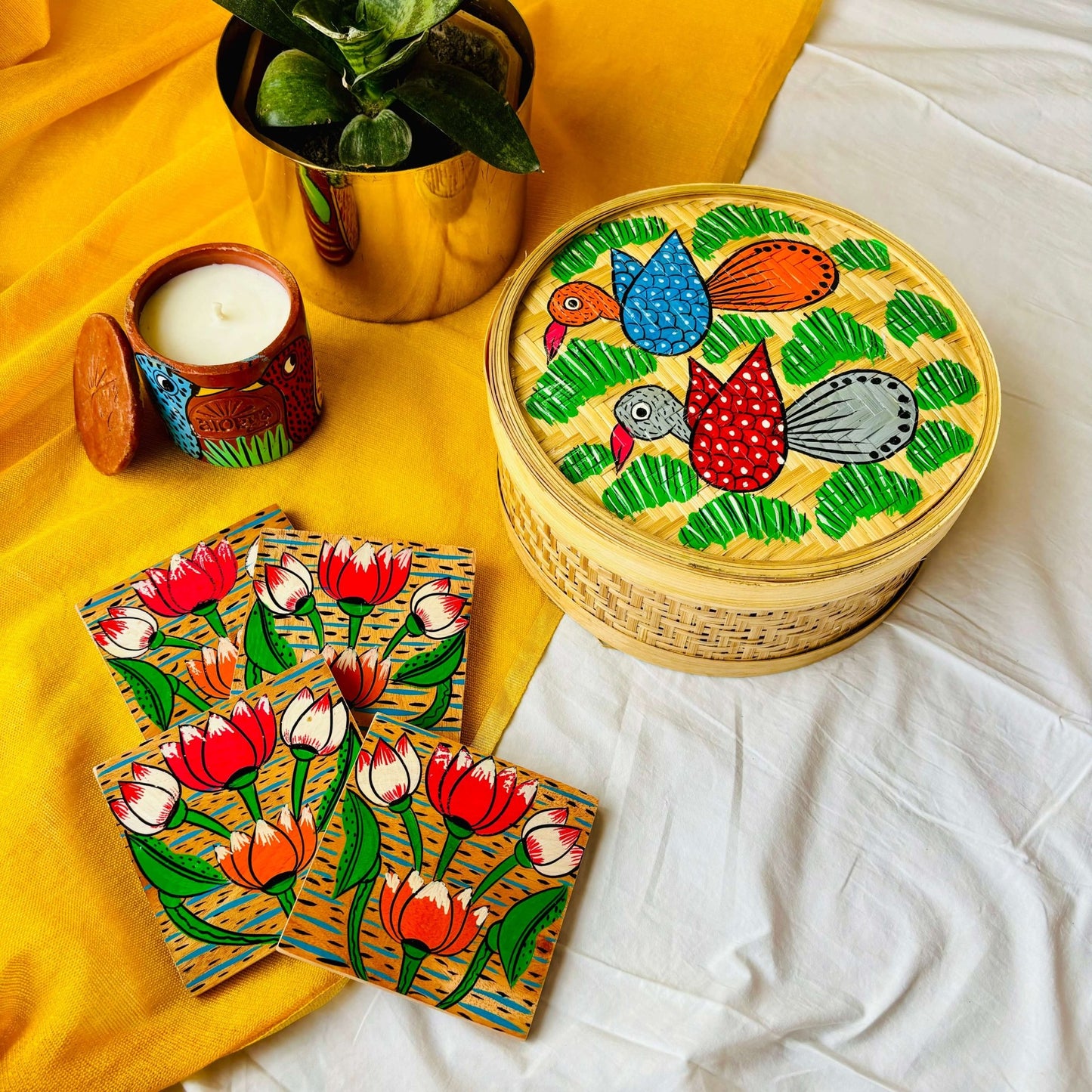 One handwoven bamboo fruit box with a motif of two birds flying over a field, a set of four square wood coasters with a motif of lotus and a double wick scented candle with a bird motif displaced against a yellow background with a plant pot beside them
