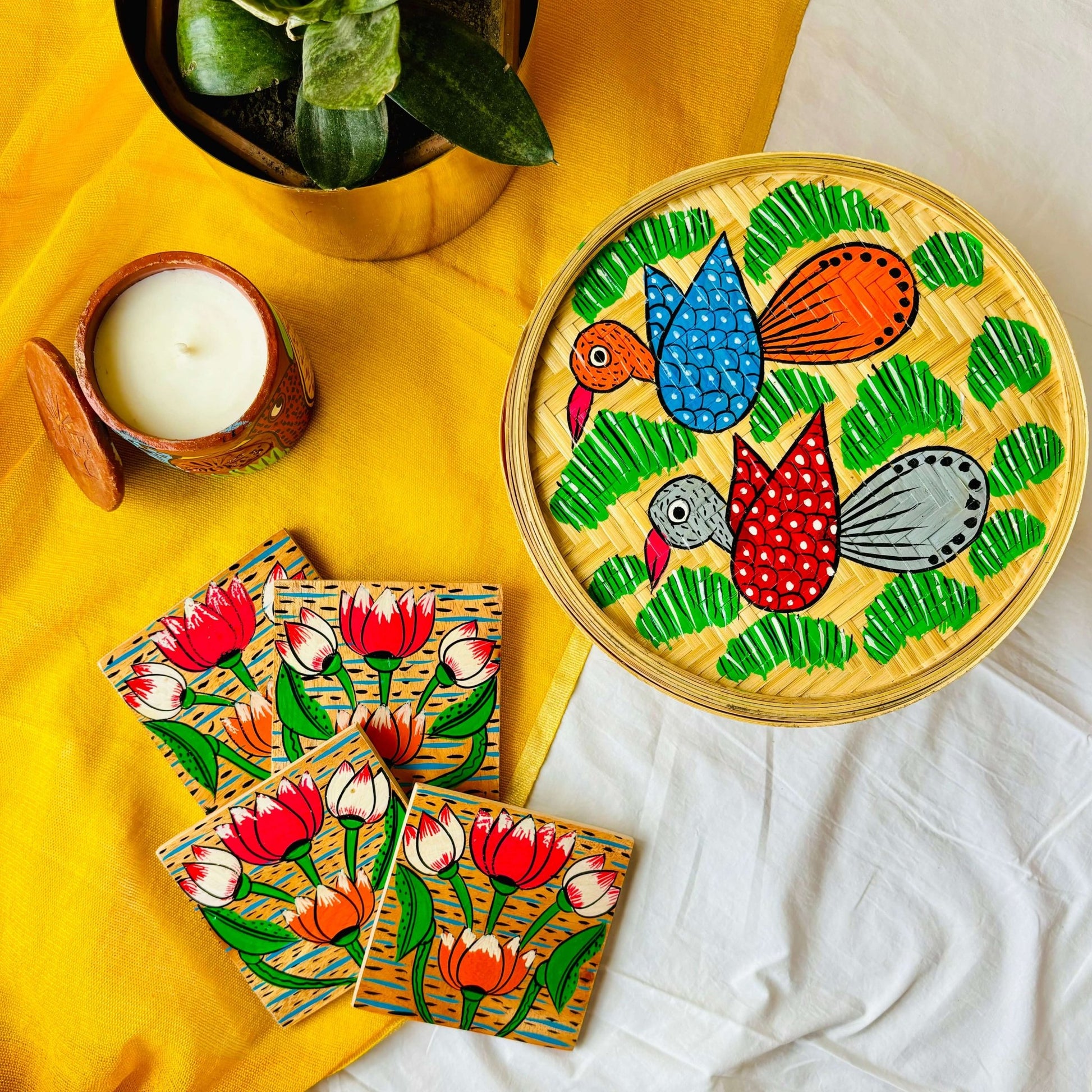 One handwoven bamboo fruit box with a motif of two birds flying over a field, a set of four square wood coasters with a motif of lotus and a double-wick scented candle displaced against a yellow background with a plant pot beside them