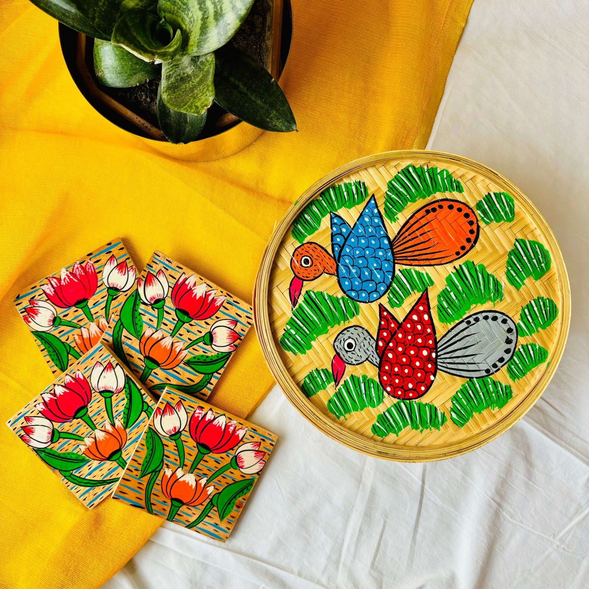 One handwoven bamboo fruit box with a motif of two birds flying over a field and a set of four square wood coasters with a motif of four lotus in each wood coaster are displaced against a yellow background with a plant pot beside them