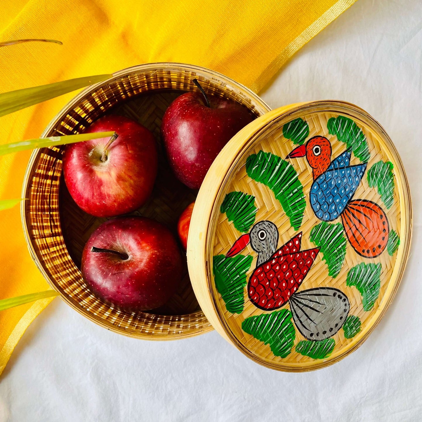 A handwoven bamboo box hand painted with one orange bird having blue wings and one gray bird having red wings flying over a green field and filled with four apples is displayed against a yellow and white background