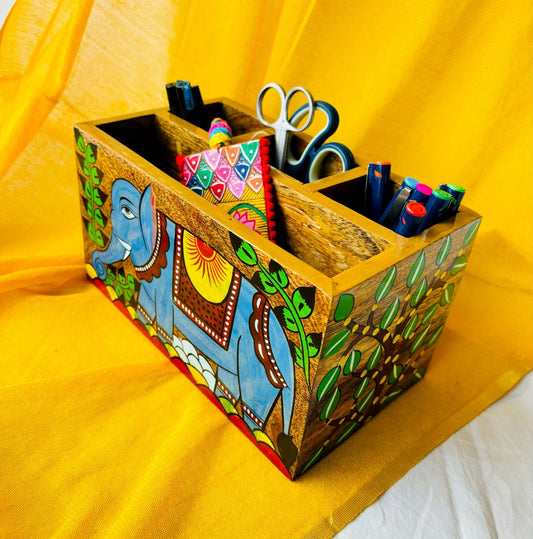 An aerial view of three pens, two scissors, six coloured pens and a bookmark placed in a 9”x5”x3” pure mango wood table organiser or wood cutlery holder wooden tissue holder with 3 small and one large compartment, handcrafted by rural artisans and painted with an elephant, tree branches and leaves motifs.