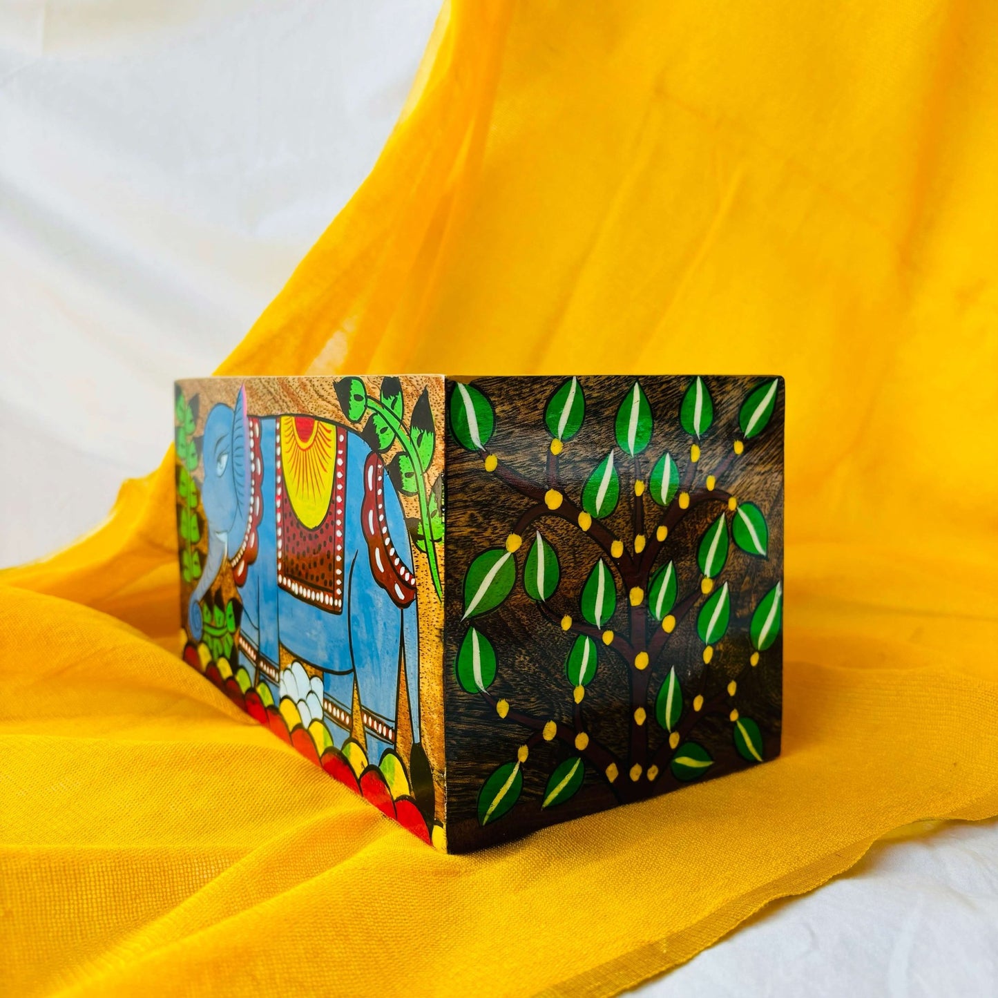 A pure mango wood table organiser or wood cutlery holder, with an elephant, tree branches and leaves hand painted on it, placed at an angle on a yellow background