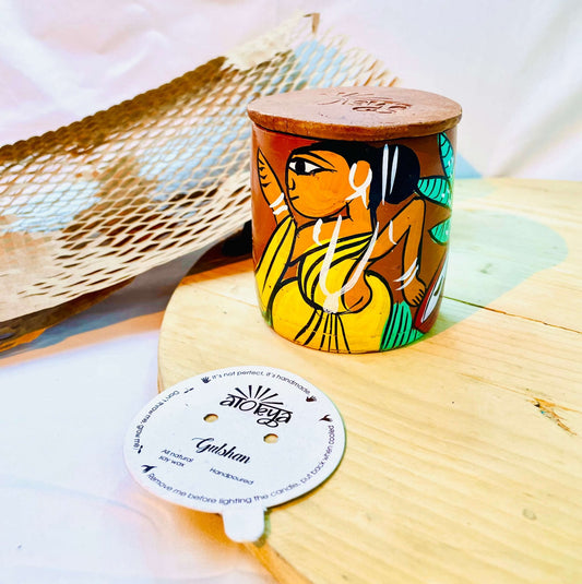 100% natural soy wax scented candle in a terracotta jar, hand painted with a tribal woman, is covered with terracotta clay candle snuffer and placed on a wooden tray near seed paper candle dust cover and honeycomb paper.