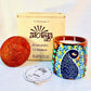 100% natural soy wax scented candle in a terracotta jar that is hand-painted with a peacock having dark blue body and light blue feathers motif. A seed paper candle dust cover is placed near the scented candle with a wooden candle box and candle snuffer in the background.