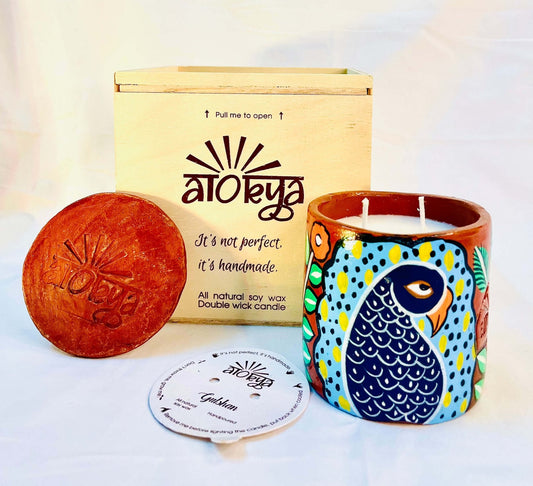 100% natural soy wax scented candle in a terracotta jar that is hand-painted with a peacock having dark blue body and light blue feathers motif. A seed paper candle dust cover is placed near the scented candle with a wooden candle box and candle snuffer in the background.