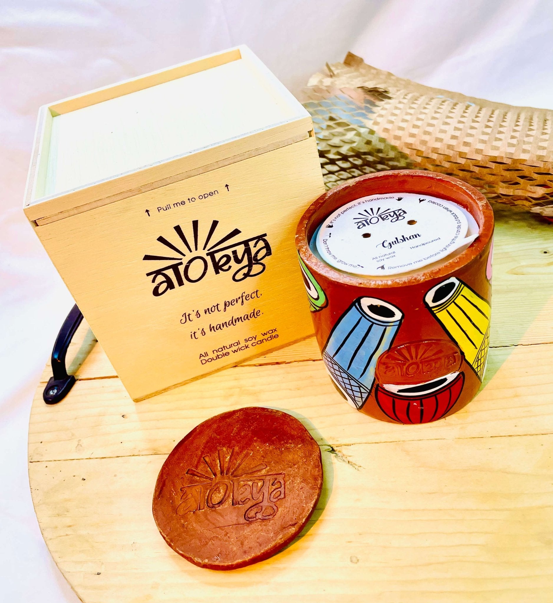 100% natural soy wax scented candle in a terracotta jar, hand painted with Indian classical musical instruments motif, and covered with seed paper candle dust cover is placed on a wooden tray. A terracotta candle snuffer is placed near the scented candle with a wooden candle box and honeycomb paper in the background.