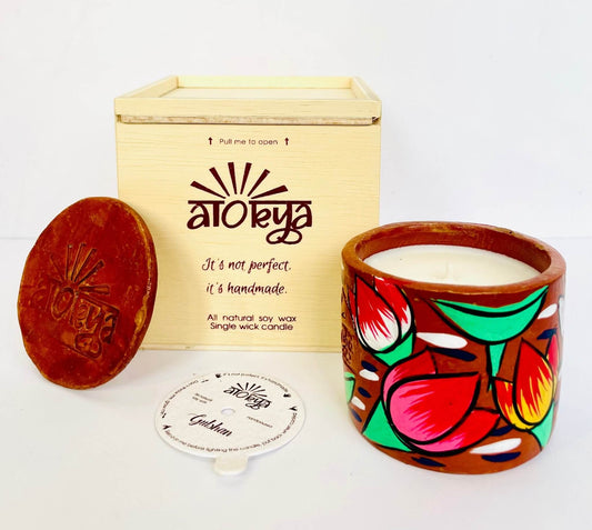 100% natural soy wax scented candle in a terracotta jar that is hand-painted with a green lotus motif. A seed paper candle dust cover is placed near the scented candle and has a wooden candle box and candle snuffer in the background.