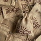 One brown jute bag with a 100% natural soy wax terracotta jar scented candle in it is placed on top of unused jute bags with alokya written on it lying flat beneath it