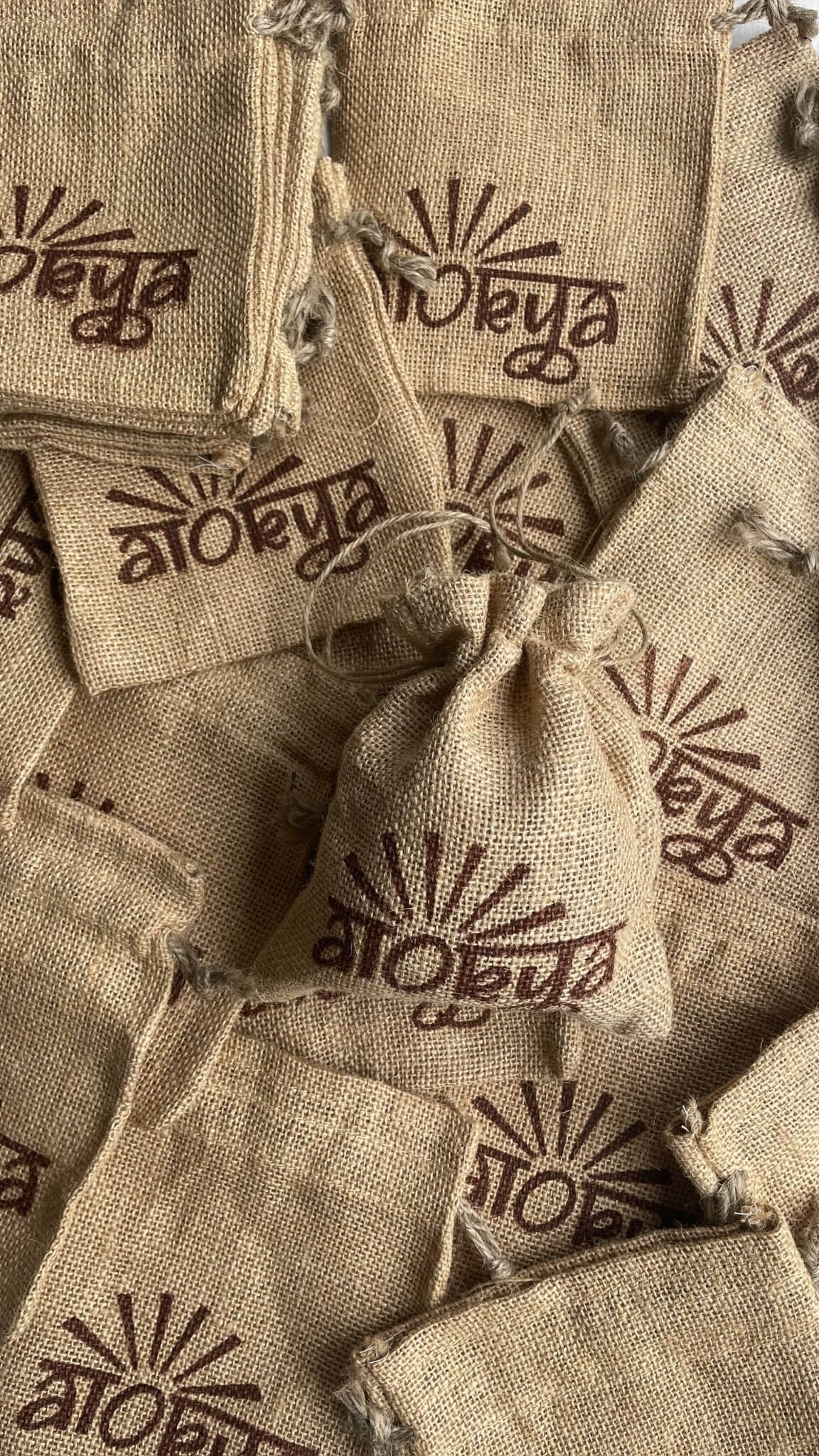 One brown jute bag with a 100% natural soy wax terracotta jar scented candle in it is placed on top of unused jute bags with alokya written on it lying flat beneath it
