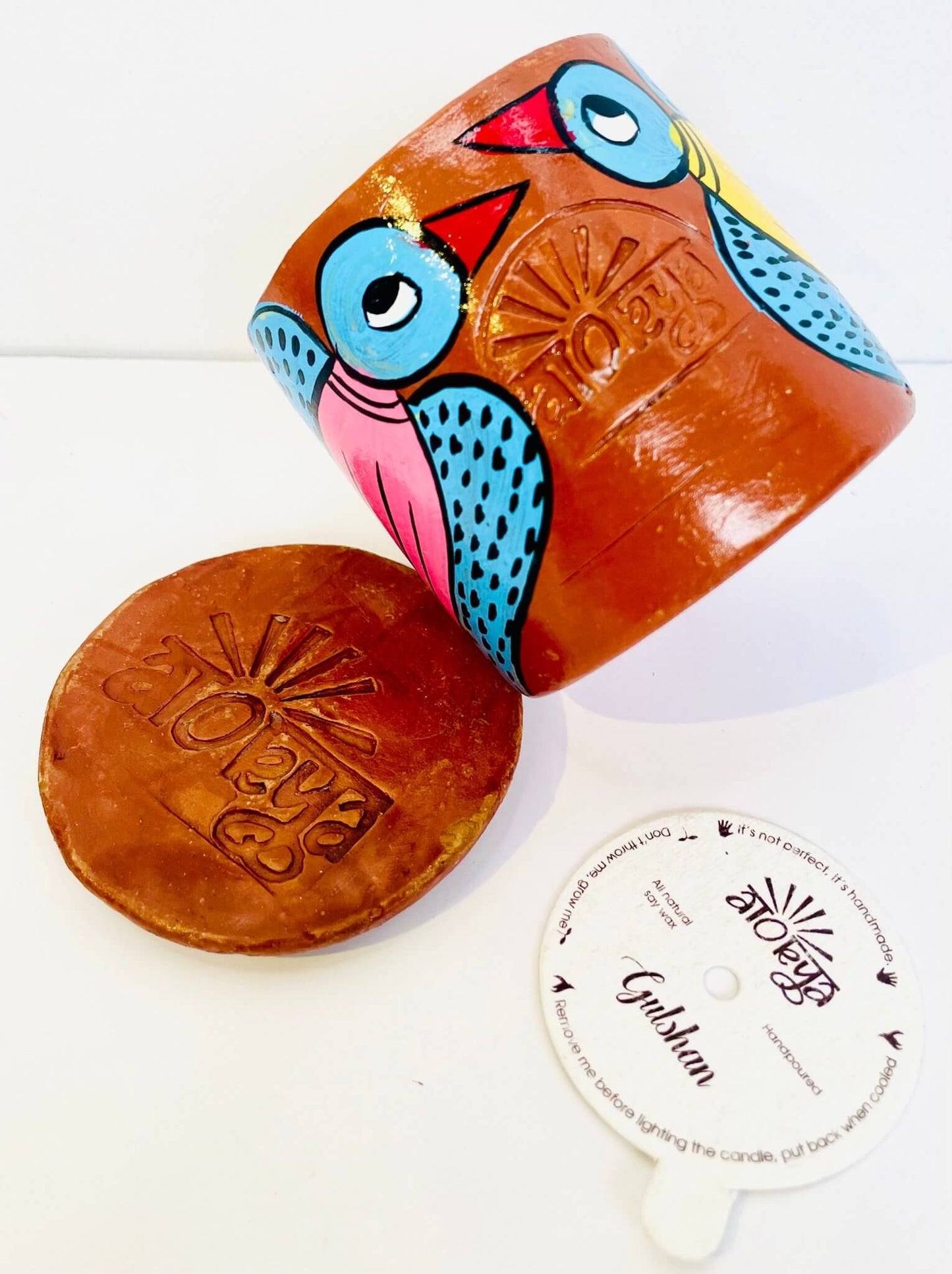 100% natural soy wax scented candle in a terracotta jar hand painted with blue birds having pink and yellow bodies is arranged together with a seed paper candle dust cover and terracotta clay candle snuffer.
