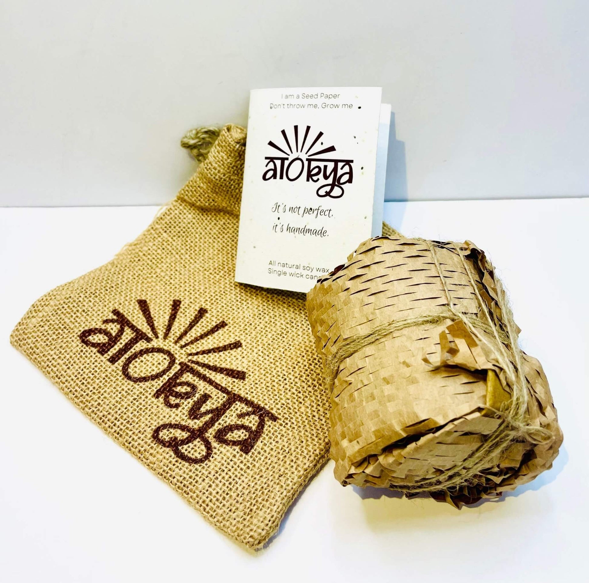 A 100% natural soy wax-scented candle wrapped in honeycomb paper is placed near a seed paper information card and jute bag.