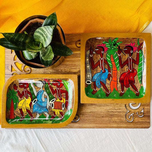 A pure mango wood square wood serving tray or trinket tray and a rectangular wooden platter hand painted with tribal characters are placed on a brown wood board near a plant pot.