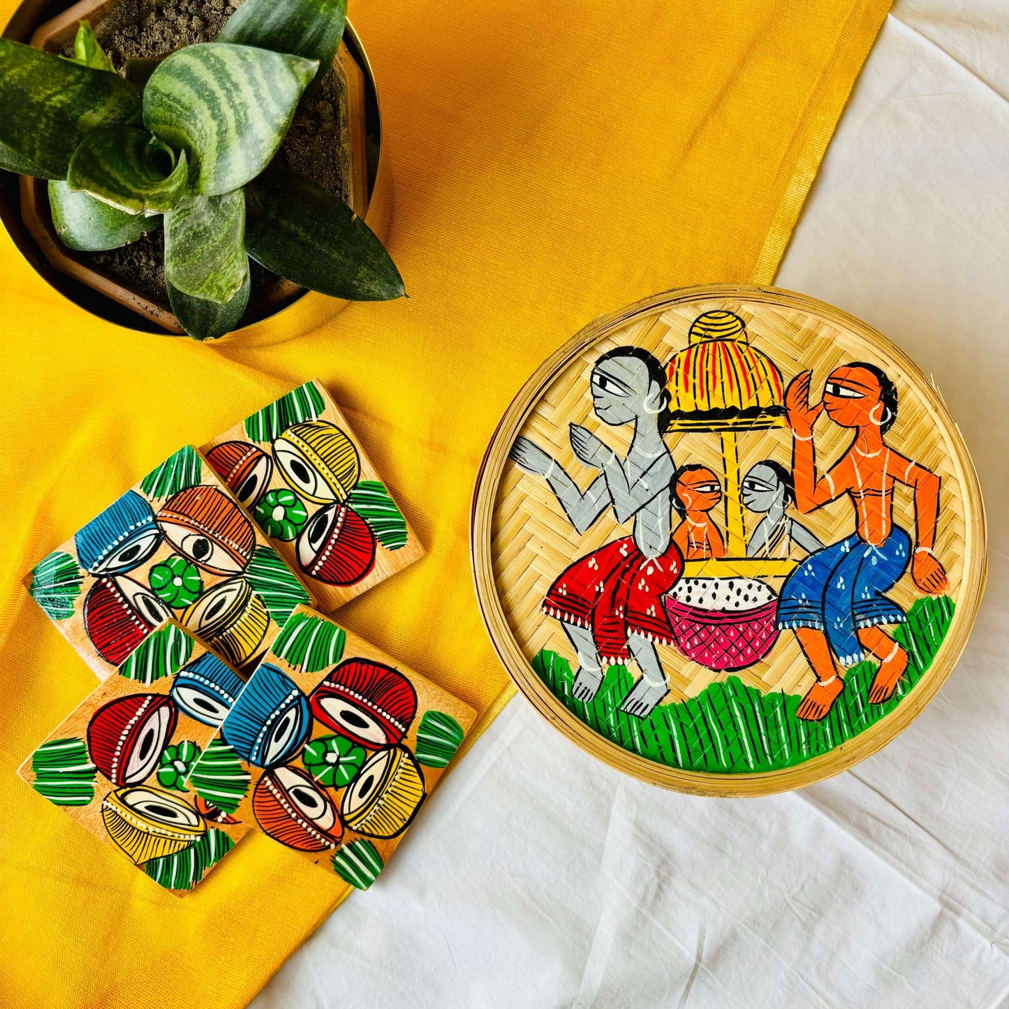 One hand-woven bamboo fruit box hand-painted with a motif of tribal characters indulged in a wedding celebration, and a set of four square wood coasters hand-painted with a motif of musical instruments are displayed against a yellow background with a plant pot beside them