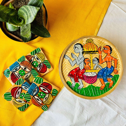 One hand-woven bamboo fruit box hand-painted with a motif of tribal characters indulged in a wedding celebration, and a set of four square wood coasters hand-painted with a motif of musical instruments are displayed against a yellow background with a plant pot beside them