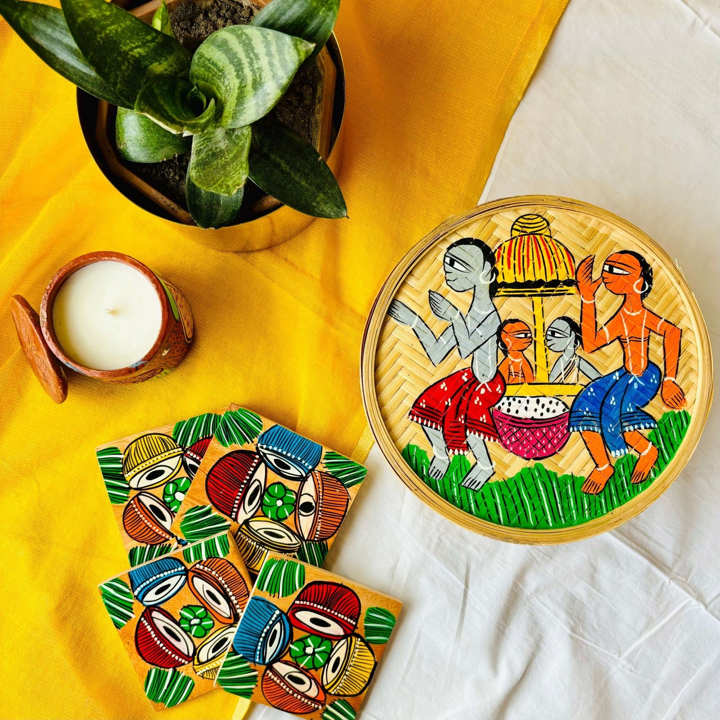 One hand-woven bamboo fruit box hand painted with a motif of tribal characters indulged in a wedding celebration, a set of four square wood coaster hand painted with a motif of musical instruments, and a double wick scented candle, all displayed against a yellow background with a plant pot beside them