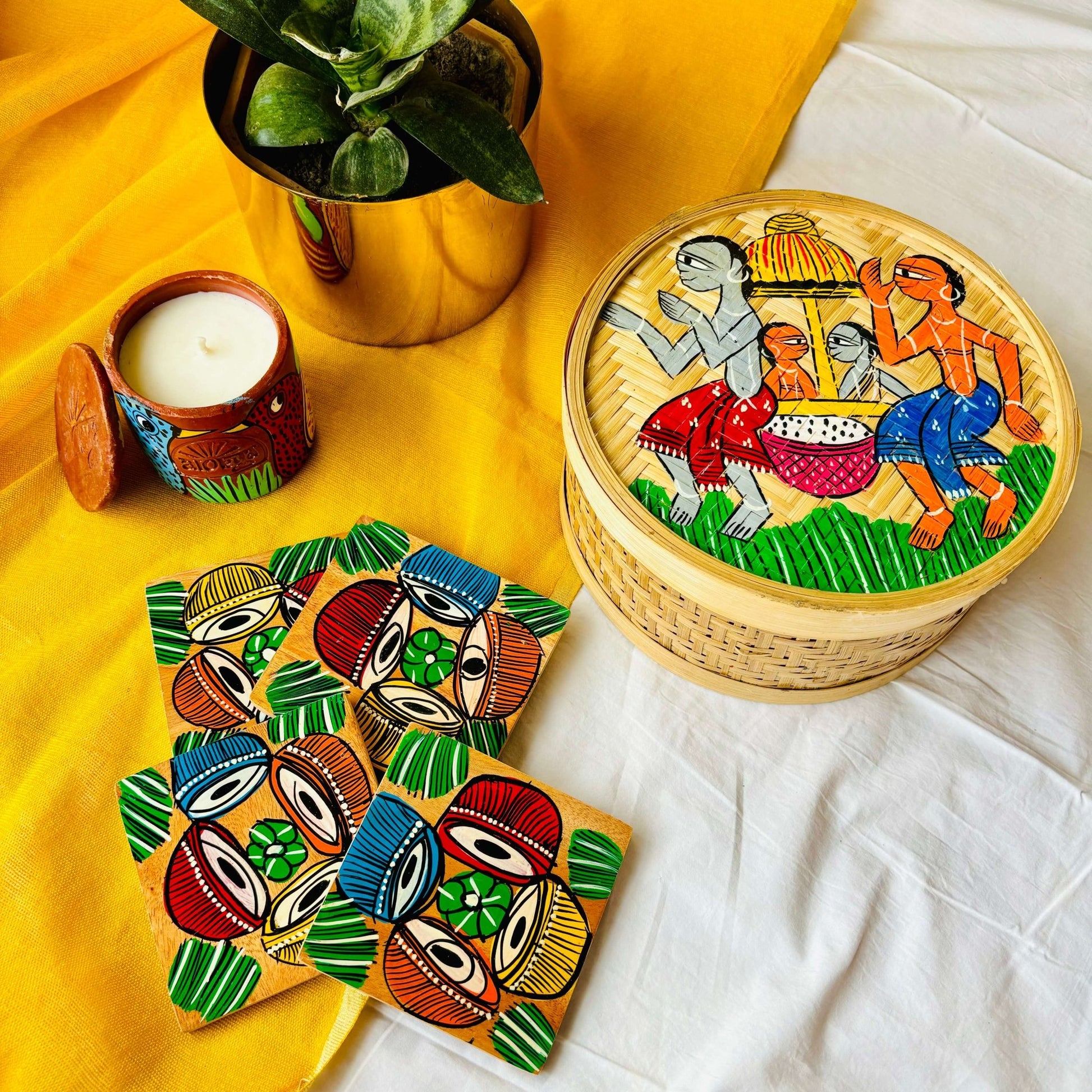One hand-woven bamboo fruit box hand painted with a motif of tribal characters indulged in a wedding celebration, a set of four square wood coasters hand painted with a motif of musical instrument, and a double wick scented candle, all displayed against a yellow background with a plant pot beside them