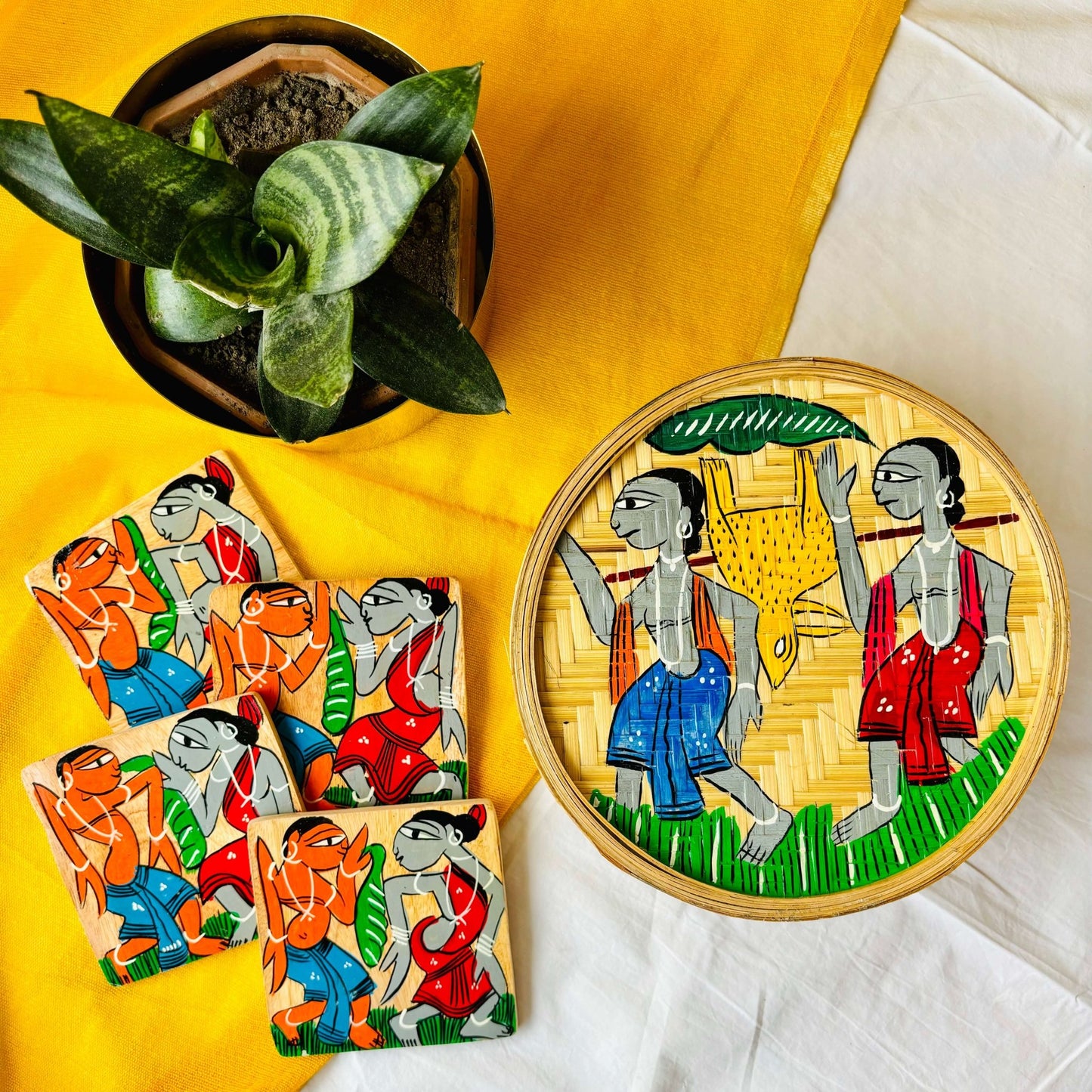 One hand-woven bamboo fruit box, and a set of four square wood coasters all hand-painted with a motif of tribal characters are displayed against a yellow background with a plant pot beside them