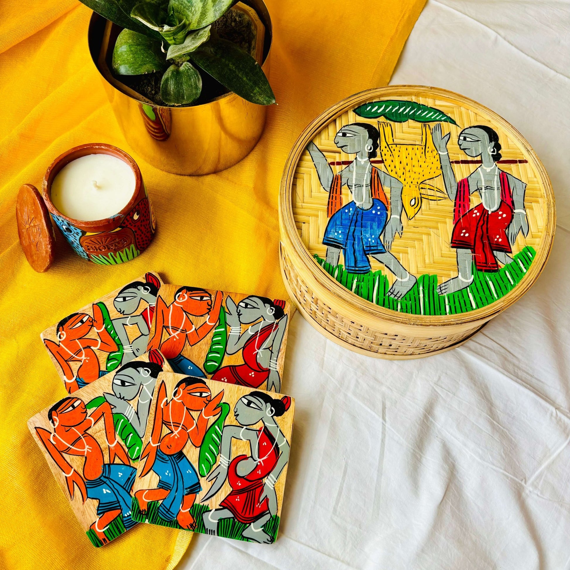 One hand-woven bamboo fruit box, a set of four square wood coaster hand painted with a motif of tribal characters, and a double wick scented candle, displayed against a yellow background with a plant pot beside them