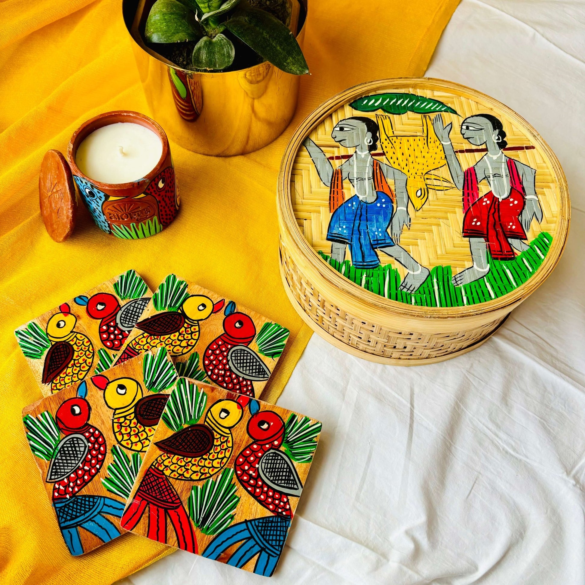 One hand-woven bamboo fruit box hand painted with a motif of tribal characters, a set of four square wood coasters hand painted with a motif of two birds on each coaster, and a double wick scented candle, displayed against a yellow background with a plant pot beside them