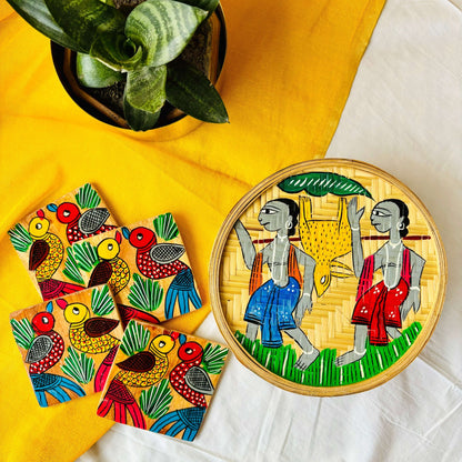 One hand-woven bamboo fruit box hand painted with a motif of tribal characters, and a set of four square wood coaster hand painted with a motif of two birds on each coaster all are displayed against a yellow background with a plant pot beside them