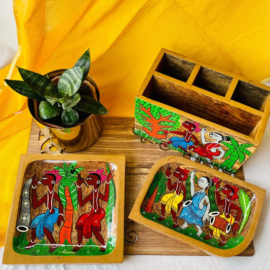 A pure mango wood square wood serving tray or trinket tray, a rectangular wooden platter and a wood cutlery holder hand painted with tribal characters are placed on a brown wood board near a plant pot.