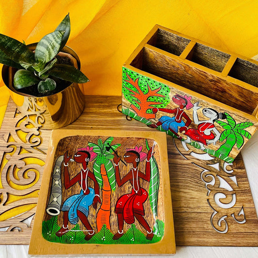 A pure mango wood square wood serving tray or trinket tray and a wood multipurpose holder or cutlery holder hand painted with tribal characters are placed on a brown wood board near a plant pot.