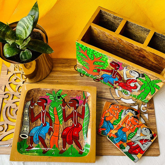 A pure mango wood square wood serving tray or trinket tray, two square wood coasters and a wood multipurpose holder or cutlery holder hand painted with tribal characters are placed on a brown wood board near a plant pot.