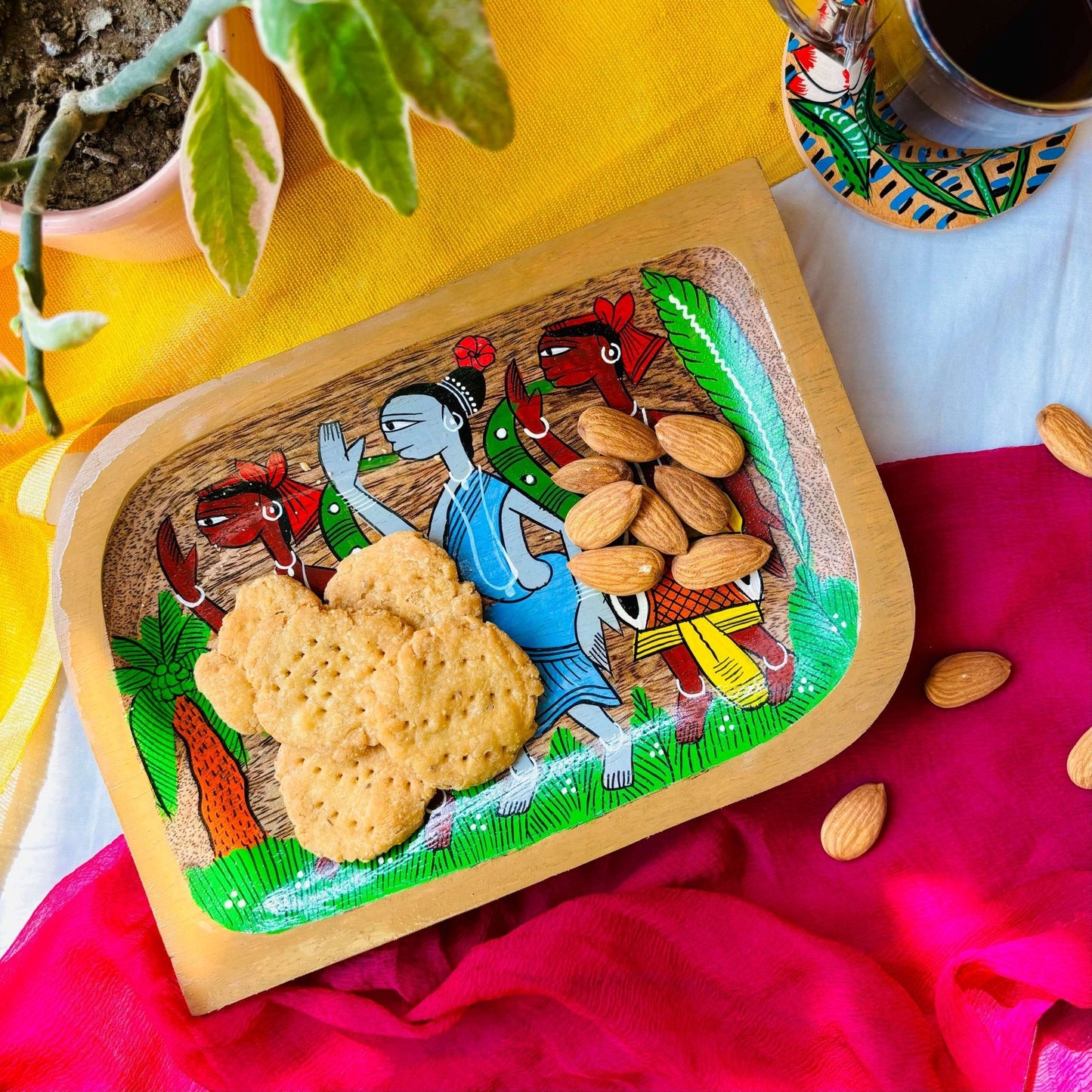 Biscuits and almonds served on a pure mango wood square wood platter/trinket tray hand painted with two tribal men wearing yellow clothes and a tribal woman wearing a blue dress dancing in a field having a coconut tree
