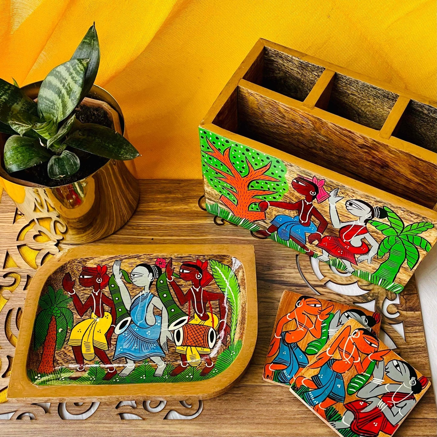 A pure mango wood rectangular wood serving tray or trinket tray, two square wood coasters and a wood multipurpose holder or cutlery holder hand painted with tribal characters are placed on a brown wood board near a plant pot.