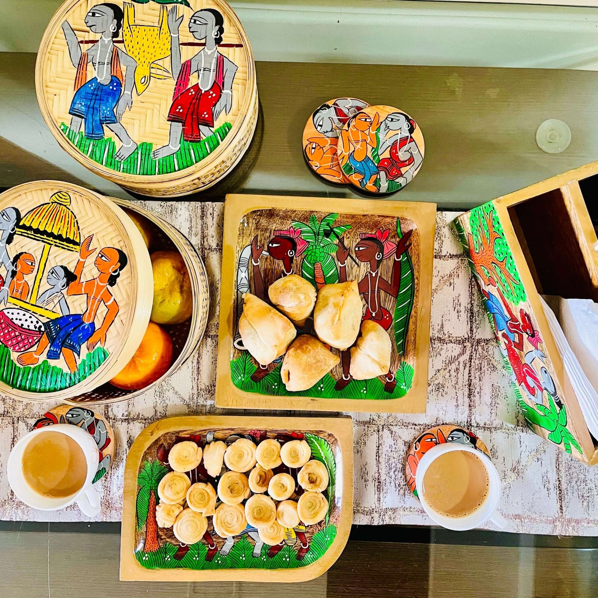 One rectangular wooden platter filled with snacks, one pure mango wood square wood serving tray filled with samosas arranged on a dining table along with four round wood coasters, two bamboo fruit boxes, one wood cutlery holder, and tea cups; all having tribal characters painted on it