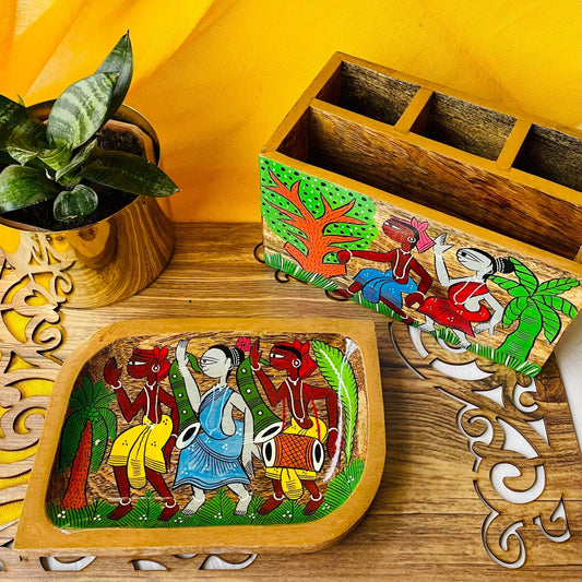 A pure mango wood rectangular wooden platter and a wood cutlery holder hand painted with tribal characters are placed on a brown wood board near a plant pot.