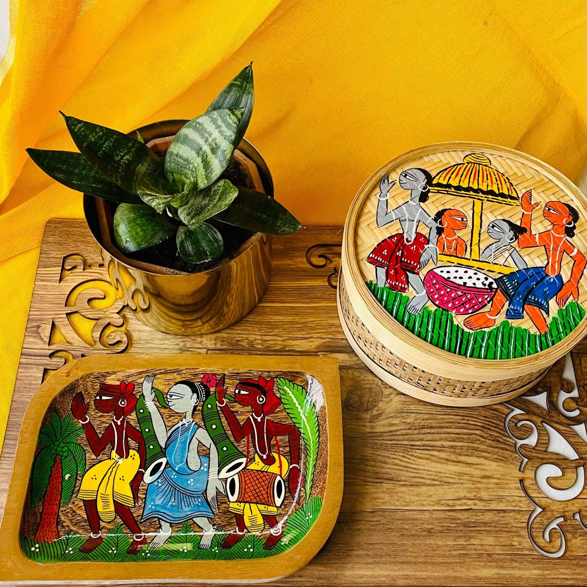 A pure mango wood rectangular wooden platter and a hand-woven bamboo utility or fruit box hand-painted with tribal characters are placed on a brown wood board near a plant pot.
