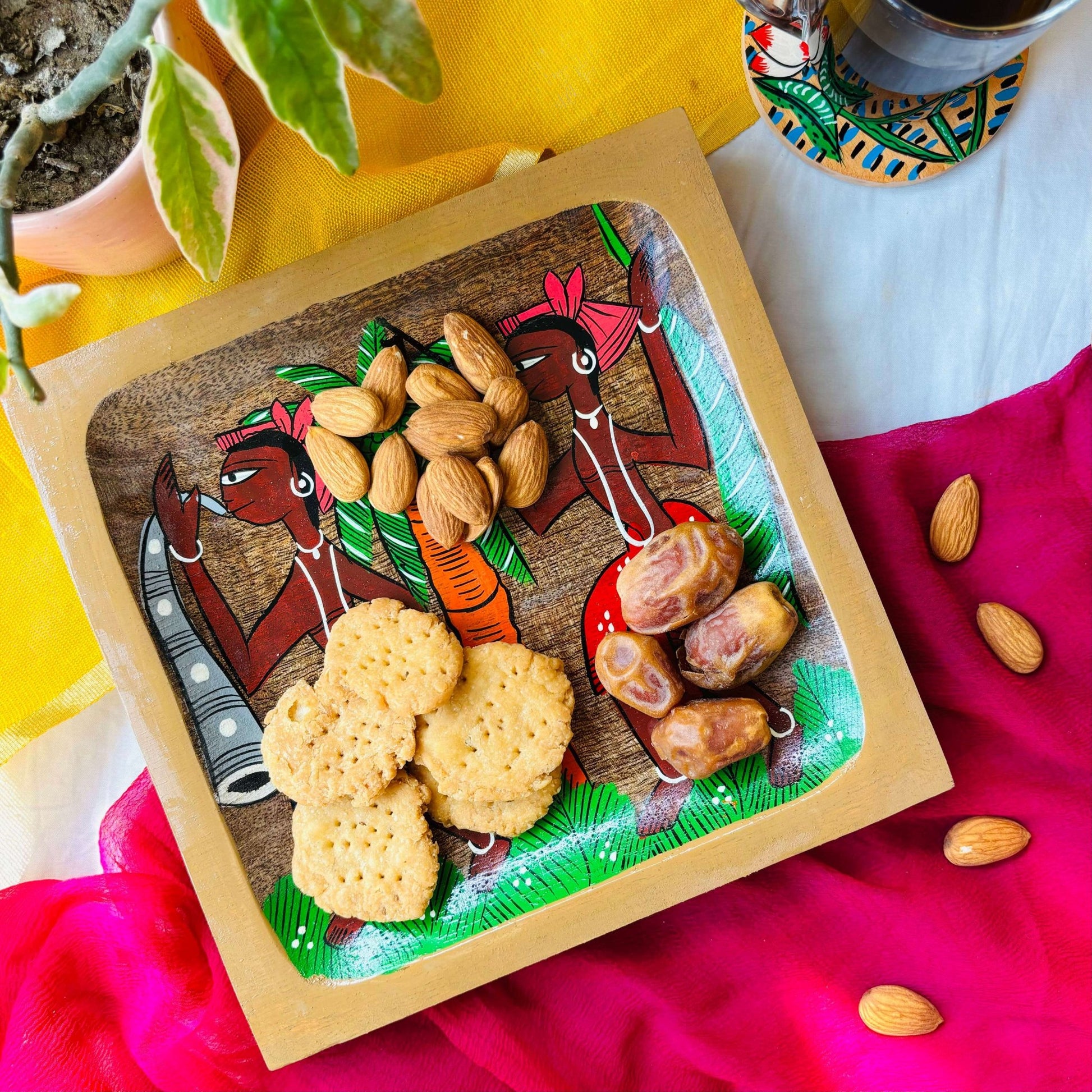 Biscuits, dates and almonds served on a pure mango wood square wood platter/trinket tray hand painted with two tribal men wearing blue and orange clothes and a coconut tree motif is displayed against a yellow and pink background