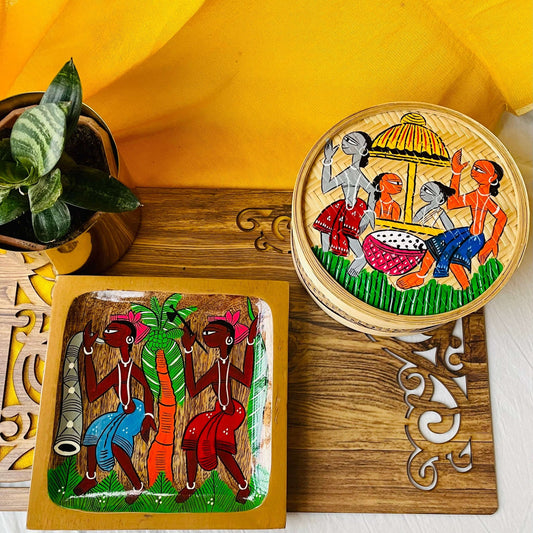 A pure mango wood square wooden platter and a hand-woven bamboo utility or fruit box hand-painted with tribal characters are placed on a brown wood board near a plant pot.