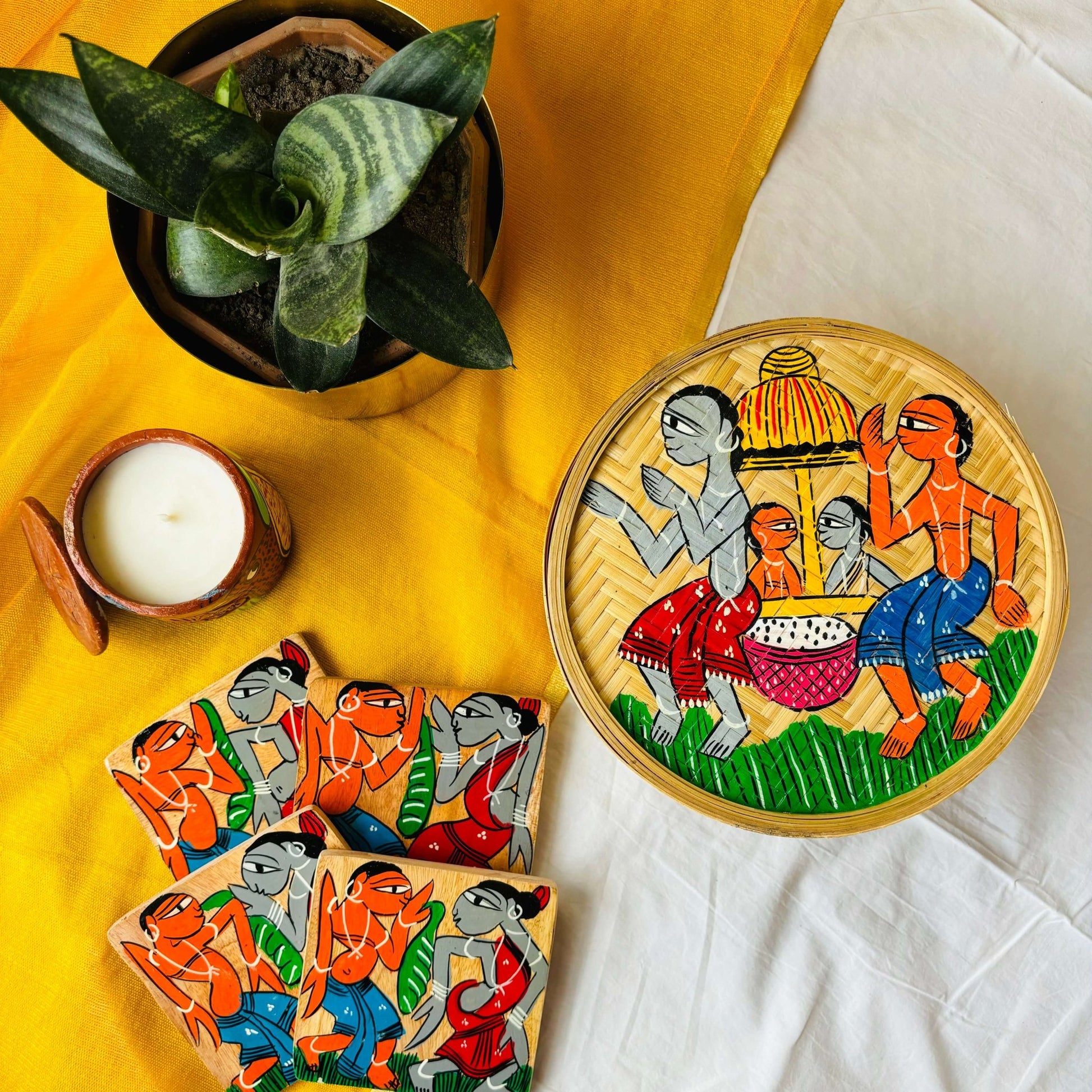 One hand-woven bamboo fruit box, a set of four square wood coasters, and a double wick scented candles, all hand painted with a motif of tribal characters indulged in a wedding celebration displayed against a yellow background with a plant pot beside them