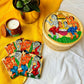 One hand-woven bamboo fruit box, a set of four square wood coasters, and a double wick scented candle, all hand painted with a motif of tribal characters indulged in a wedding celebration displayed against a yellow background with a plant pot beside them