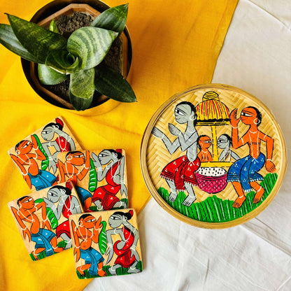 One hand-woven bamboo fruit box, and a set of four square wood coasters all hand-painted with a motif of tribal characters indulged in a wedding celebration are displayed against a yellow background with a plant pot beside them
