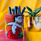 Six coloured pens organised in a terracotta pen stand hand painted with a painting of a tribal man placed on a book in front of a plant pot