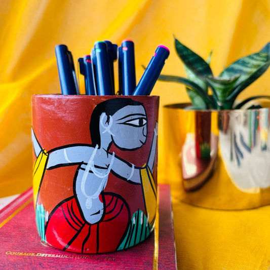 Six coloured pens organised in a terracotta pen stand hand painted with a painting of a tribal man placed on a book in front of a plant pot