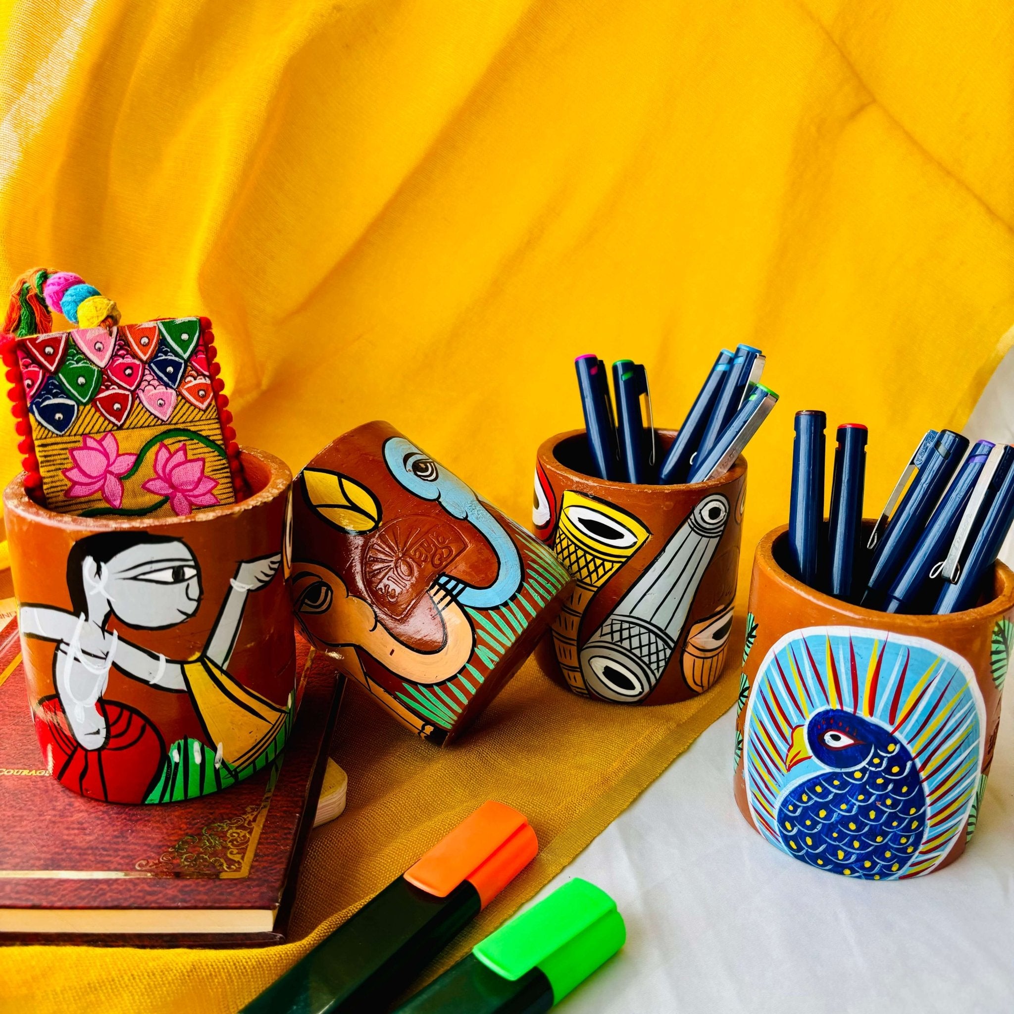 Two terracotta pen stands holding six coloured pens each hand-painted with peacock and musical instruments, one elephant-painted terracotta pen stand and a terracotta pen holder painted with a motif of tribal man are placed against a yellow background.