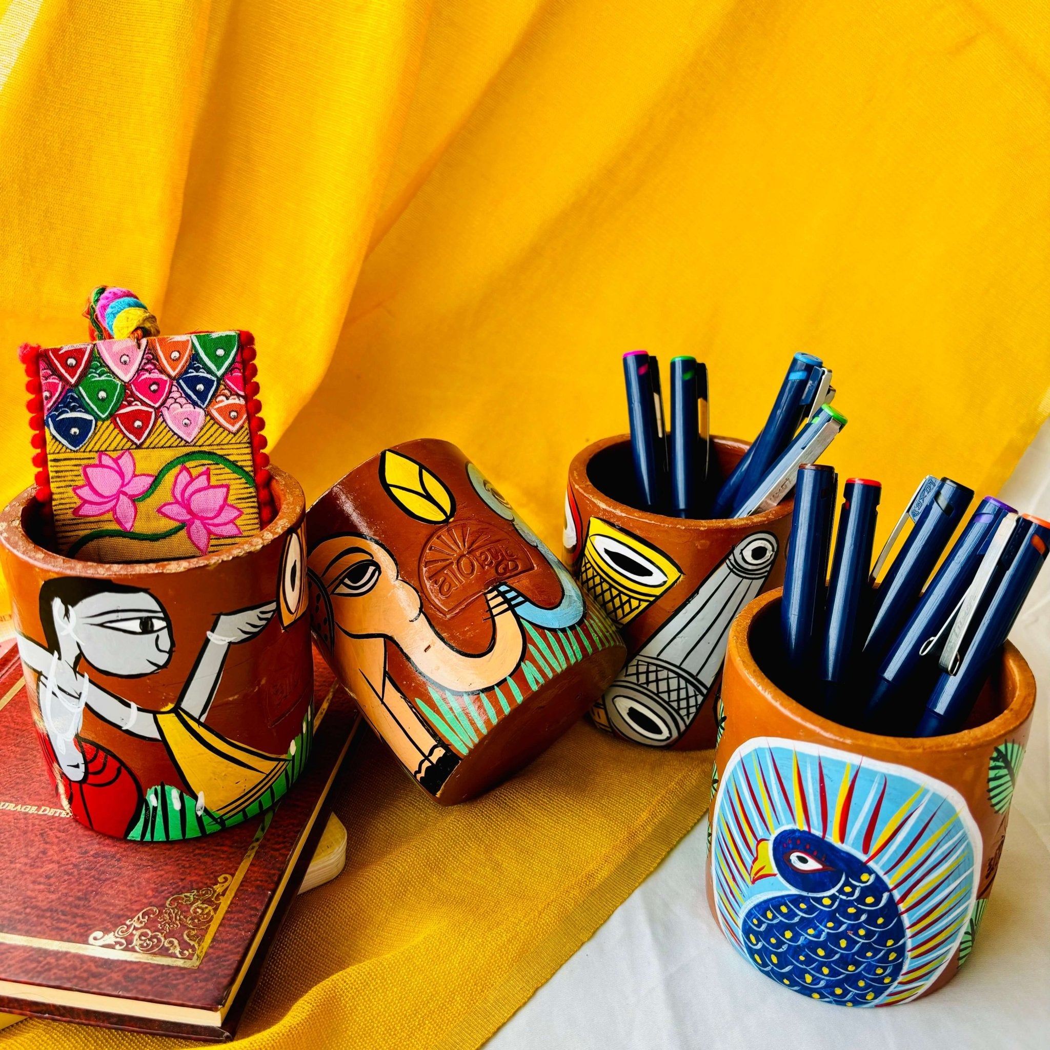 Two terracotta pen stands holding six coloured pens each hand-painted with peacock and musical instruments, one elephant-painted terracotta pen stand and a terracotta pen holder painted with a motif of a tribal man is placed against a yellow background.