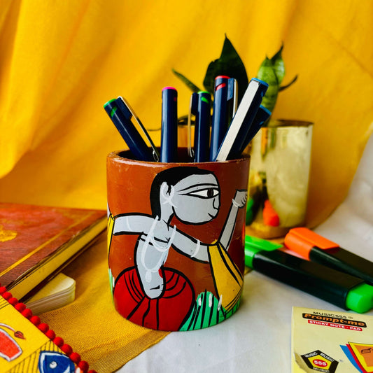 Six coloured pens organised in a terracotta pen stand hand painted with a tribal man dancing are placed against a yellow background along with green and orange highlighter, sticky notes, a book and a plant pot.