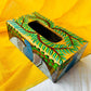 An aerial view of a 9”x5”x3” pure mango wood wooden tissue holder, handcrafted by rural artisans and painted with peacock, tree branches and leaves motifs