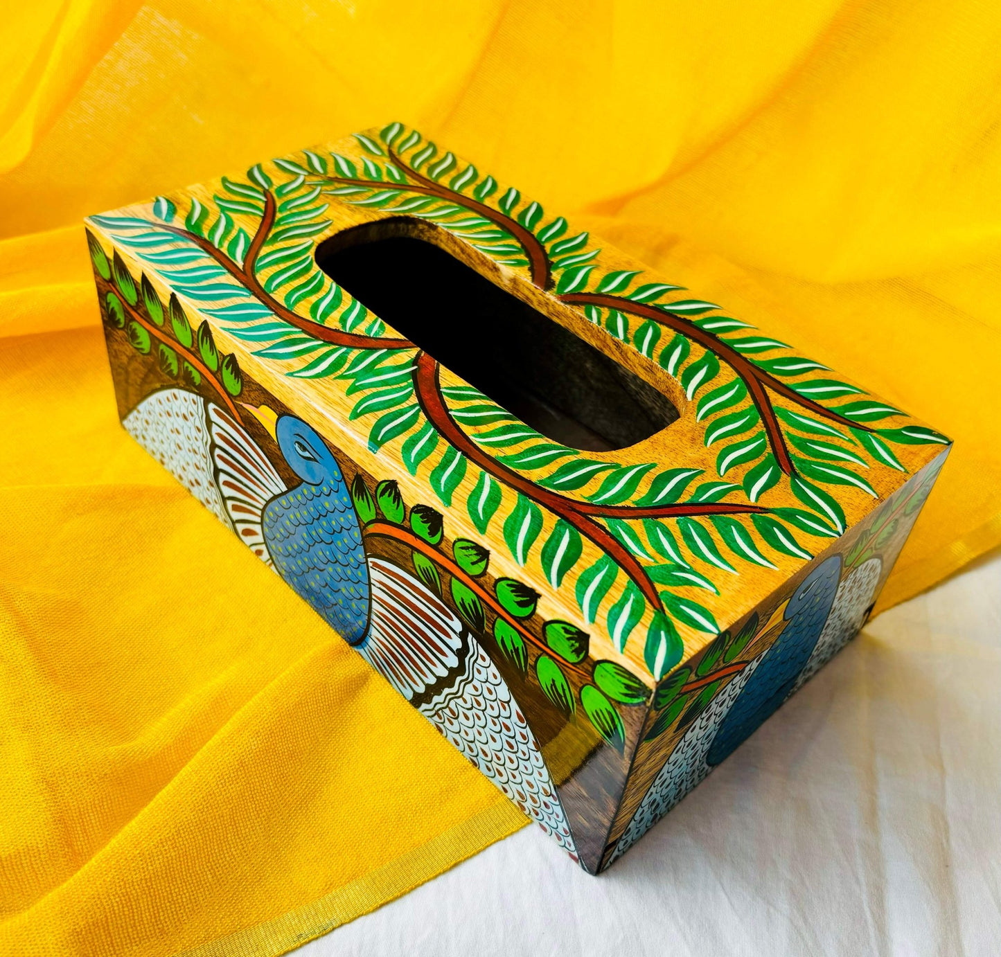 An aerial view of a 9”x5”x3” pure mango wood wooden tissue holder, handcrafted by rural artisans and painted with peacock, tree branches and leaves motifs