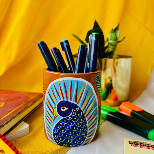 Six coloured pens organised in a terracotta pen stand hand painted with a motif of a blue peacock placed against a yellow background along with green and orange highlighter, sticky notes, a book and a plant pot.