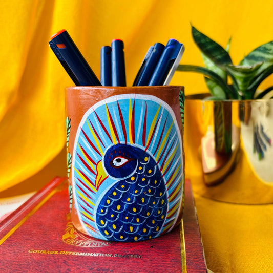 Six coloured pens organised in a terracotta pen stand hand painted with a painting of a blue peacock placed in front of a plant pot