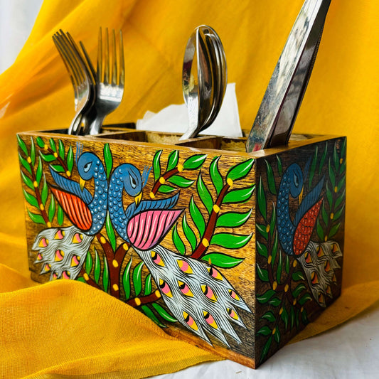 A pure mango wood table organiser or wood cutlery holder, with two peacocks, tree branches and leaves paintings placed at an angle with a yellow background and three spoons, three forks and knives organised in it