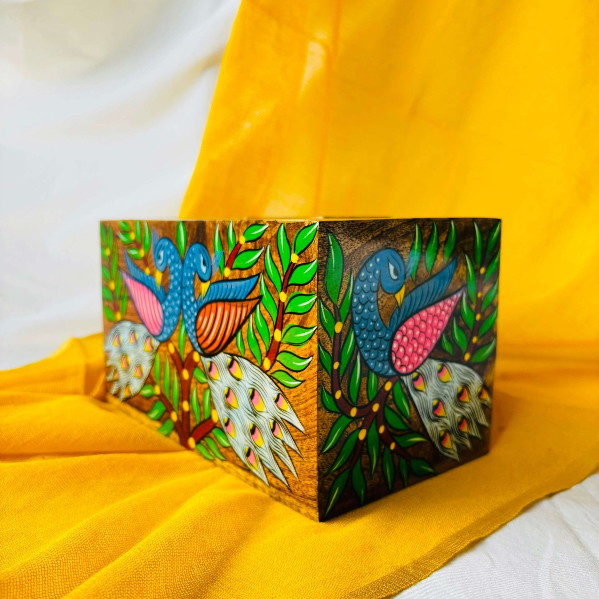 A pure mango wood table organiser or wood cutlery holder, with two peacocks, tree branches and leaves hand painted on it, placed at an angle on a yellow background