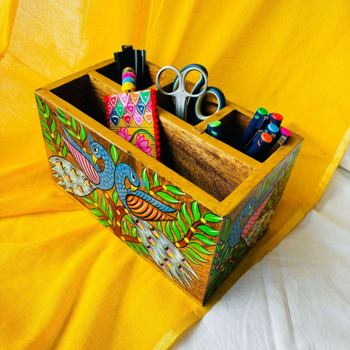 An aerial view of three pens, two scissors, six coloured pens and a bookmark placed in a 9”x5”x3” pure mango wood table organiser or wood cutlery holder wooden tissue holder with 3 small and one large compartment, handcrafted by rural artisans and painted with two peacocks, tree branches and leaves motifs